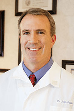 The image shows a man in a professional setting, wearing a white lab coat with name tags and a stethoscope around his neck. He is smiling at the camera, posing for the photograph.