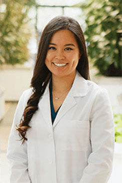 The image features a person standing in front of a plant, wearing a white lab coat and smiling.