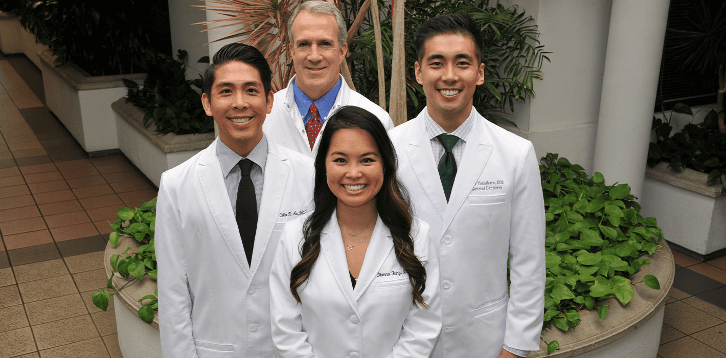 The image is a photograph of four individuals, likely medical professionals or students, posing together in front of an indoor plant. They are wearing white lab coats and stethoscopes, suggesting a healthcare setting.