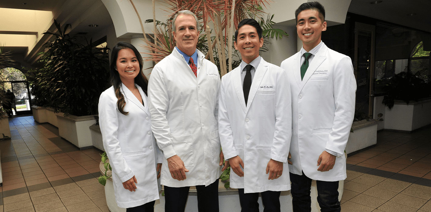 The image shows a group of four individuals, likely professionals given their attire, posing together for a photograph. They are standing in front of a potted plant and appear to be indoors with a tiled floor and a wall-mounted clock visible in the background.