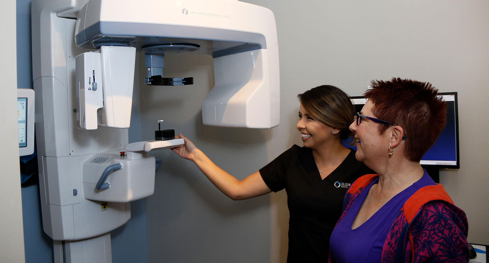 This is an image featuring two individuals, one of whom appears to be operating a dental or medical device, possibly a scanner or X-ray machine. The setting suggests a professional environment, likely a dental or medical office.