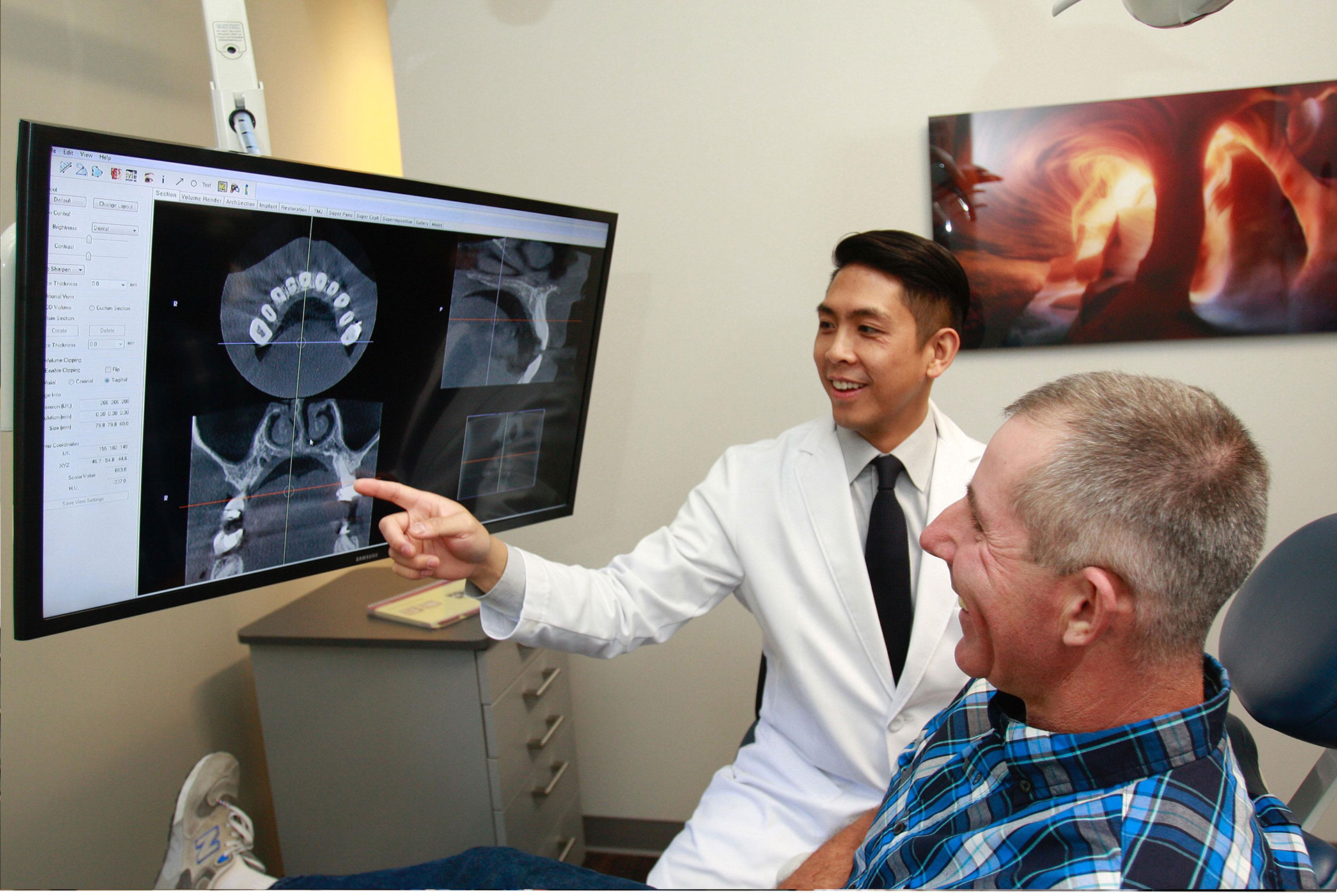 The image shows a medical professional, possibly a radiologist or dentist, using a large screen to explain an X-ray image to a patient in a dental or medical office setting.