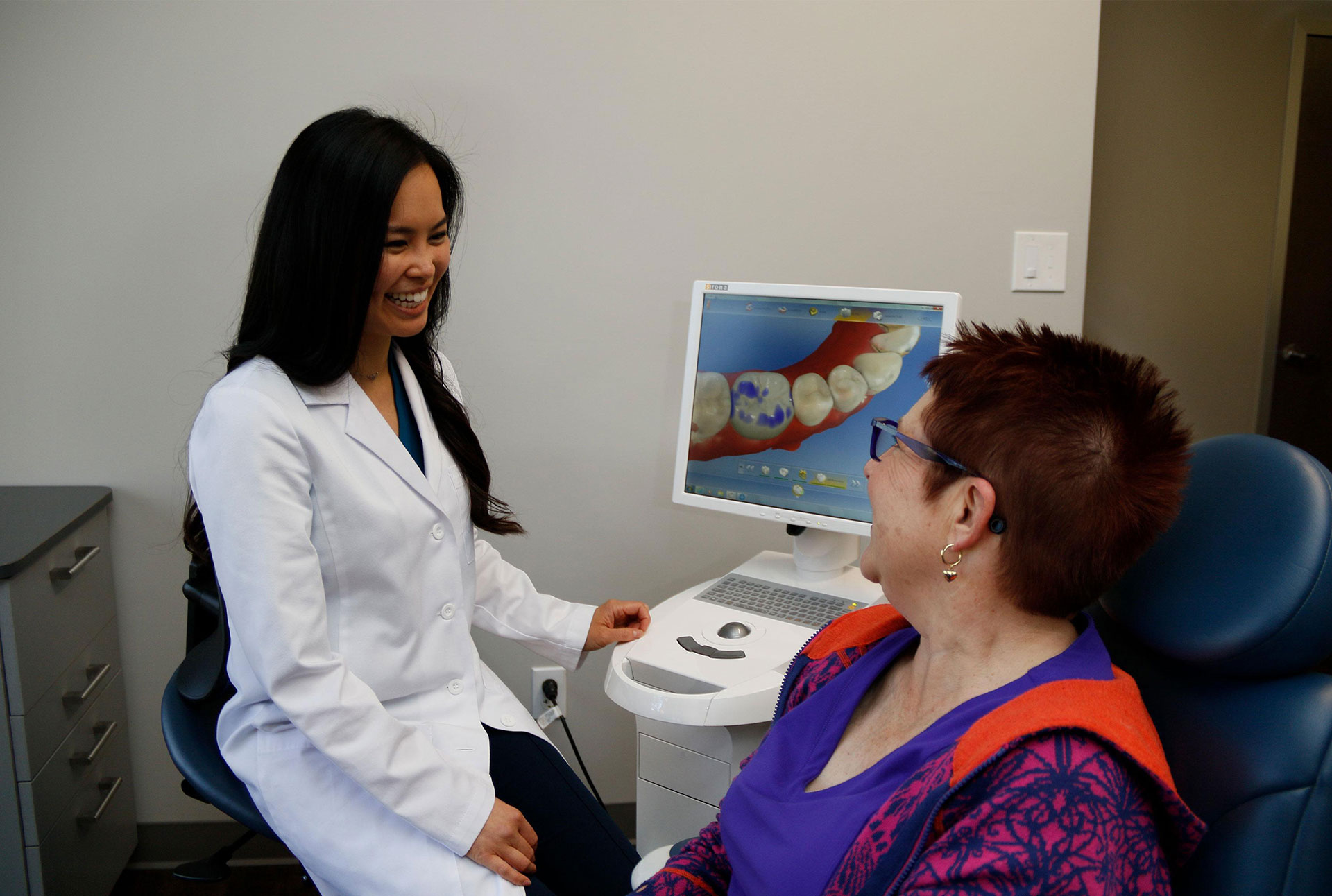 A dental professional in a white coat assisting an older woman with a toothbrush demonstration, set against a clinical background.