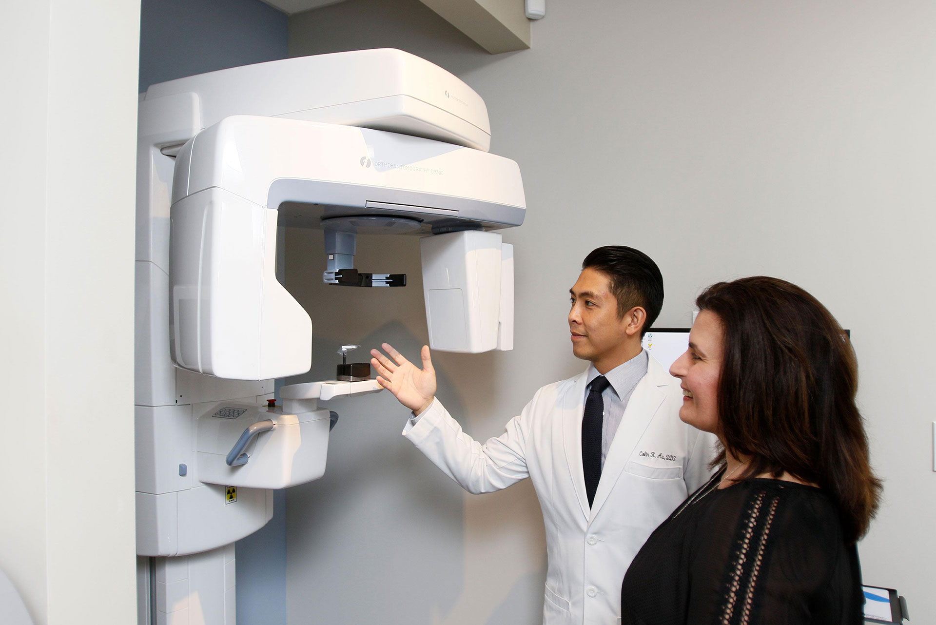 The image shows a person standing next to a large, modern-looking 3D scanner in what appears to be a medical or dental office setting. A second person is pointing at the scanner with interest.