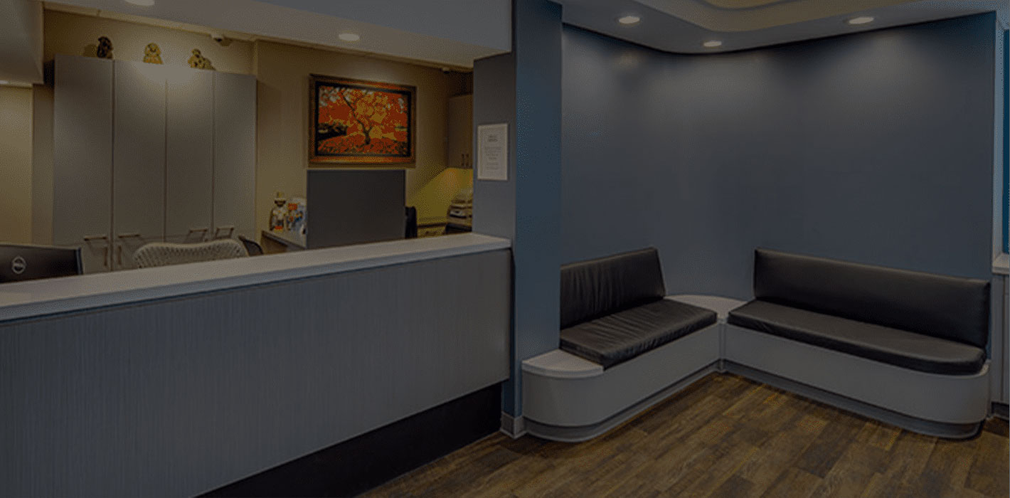 The image shows an interior space that appears to be a waiting area or lobby, featuring a reception desk with a computer monitor, a seating area with two black chairs and a wooden bench, a countertop with a sink, and a wall with a framed picture. The color scheme is neutral, with shades of gray and blue dominating the space.