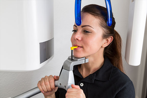 Woman in dental office holding a dental tool while wearing a blue mouthguard, with an X-ray machine behind her.