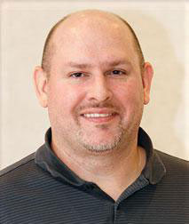 The image shows a bald man with a mustache, smiling slightly and looking directly at the camera. He is wearing a dark shirt with a collar and appears to be indoors.