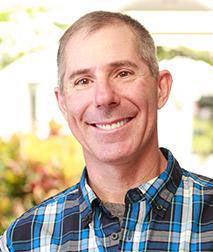 The image shows a man smiling at the camera, wearing a plaid shirt and glasses. He has short hair and appears to be indoors with a warm lighting ambiance.