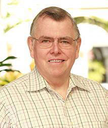 The image is a portrait of an older man with glasses, wearing a light-colored shirt and dark pants. He has short gray hair and is smiling slightly at the camera.