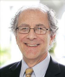 The image is a photograph of an older man with glasses, smiling at the camera. He has gray hair and appears to be wearing a suit with a tie. There s no visible text on the image.