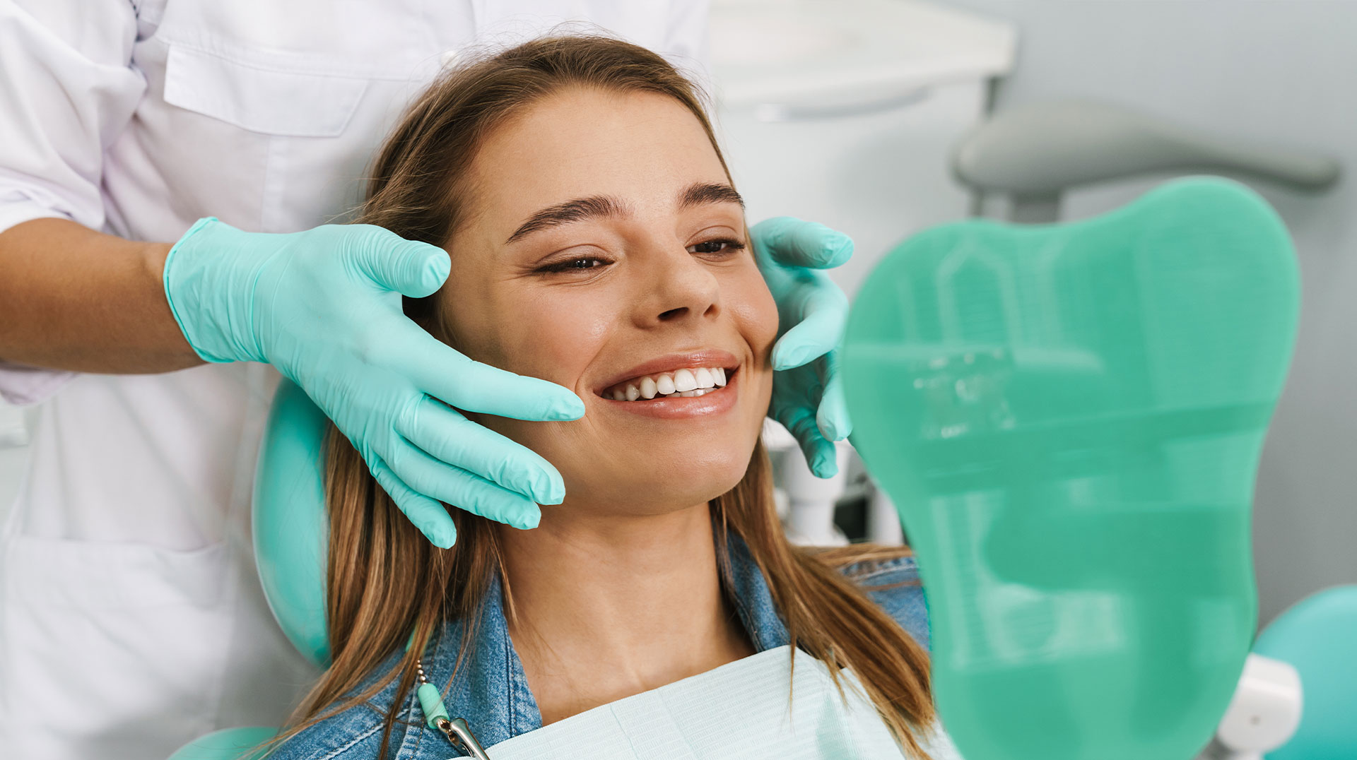 A smiling woman in a dental chair receiving a dental cleaning, with a dental hygienist working on her teeth.