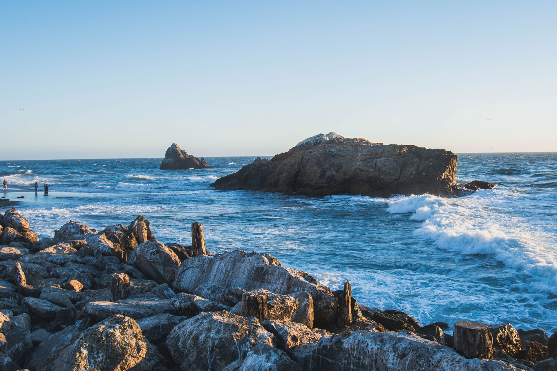 A coastal scene featuring a rocky shoreline with crashing waves, a large boulder prominently in the foreground, and a clear blue sky above.