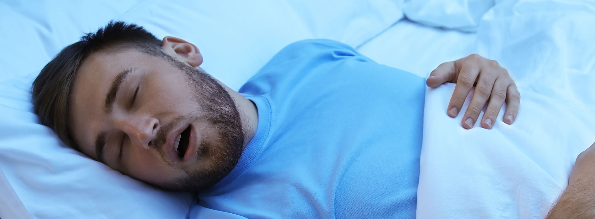 The image shows a man lying in bed with his mouth open, suggesting he is either asleep or in the midst of speaking or breathing.