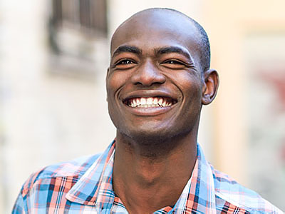 The image shows a smiling man with short hair, wearing a patterned shirt, standing outdoors against a building facade.