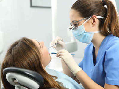 The image shows a dental professional in a white coat and face mask, using dental instruments to work on the teeth of a patient who is seated in a dental chair.
