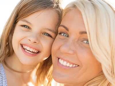 The image shows a woman and a young girl smiling at the camera, with the woman appearing to be older than the child. They are outdoors in bright sunlight, and both individuals have light skin tones.
