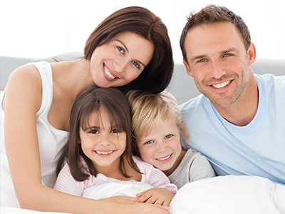 The image depicts a family of four, including an adult couple and their two young children, all sharing a moment together on a bed.