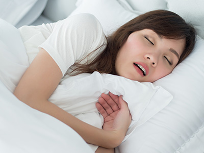 A woman in a white shirt is lying down with her eyes closed, appearing to be asleep or resting. She has a pillow under her head and is holding a blanket close to her face.