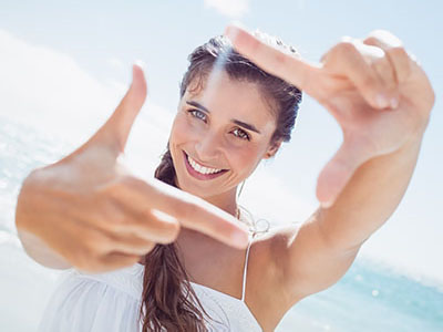 A woman with long hair, wearing a white top, is smiling and holding up two fingers in front of her face. She appears to be at the beach, as suggested by the clear sky and the ocean visible behind her.